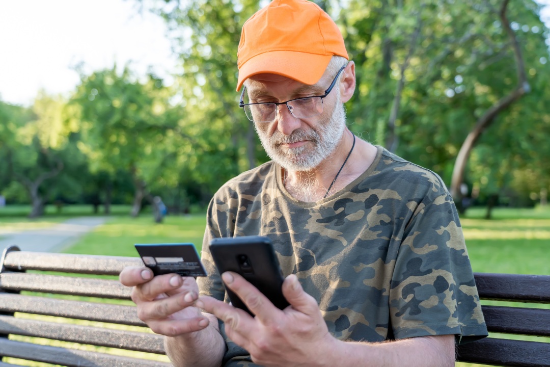 Senior man using credit bank card and smartphone while transferring money, pay debt, purchases online shopping on bench outdoors, financial virtual service