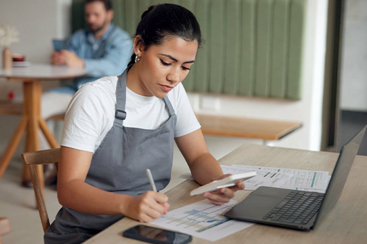 A woman in an apron works on a laptop, focused on tasks related to new tax rules for 2026.