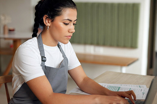 A woman in an apron works on a laptop, focused on tasks related to new tax rules for 2026.
