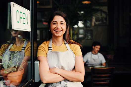 A cheerful woman wearing an apron poses in front of a restaurant, ready to greet customers with a warm smile.